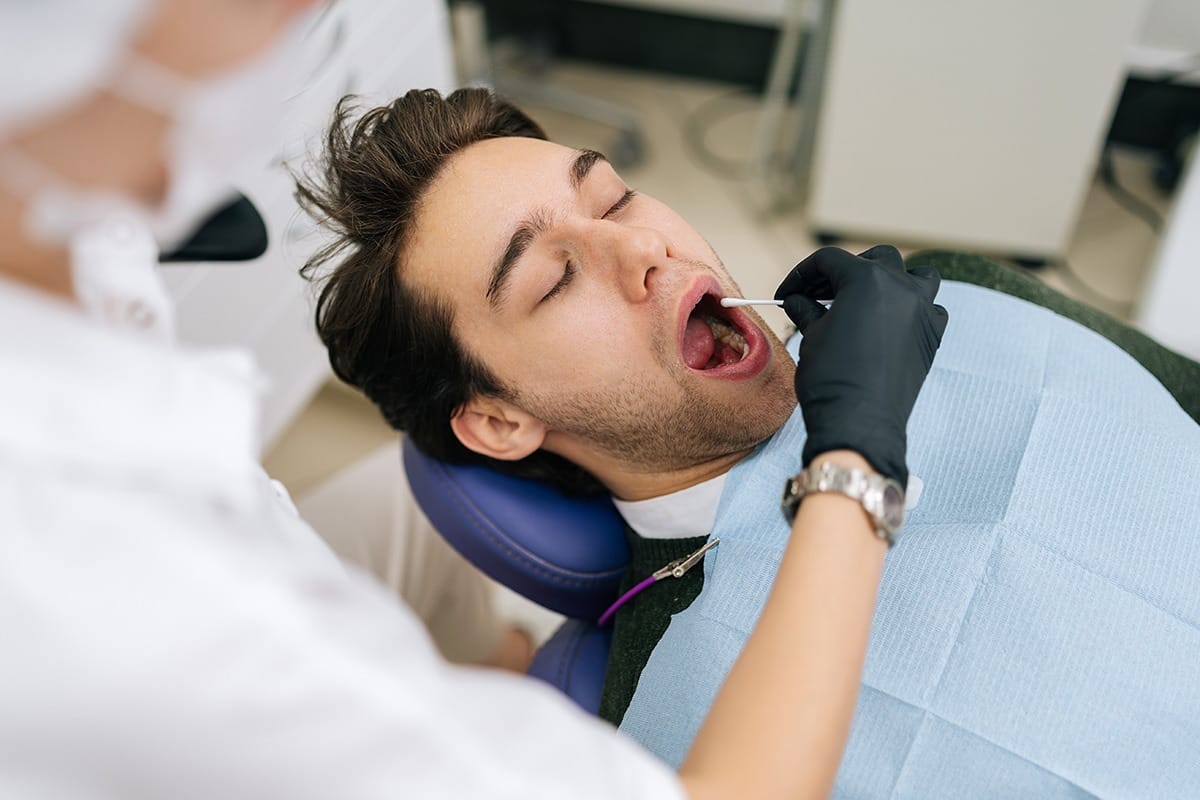 View from shoulder of unrecognizable female doctor taking saliva test from male patient mouth with cotton swab. Dentist using cotton swab to lubricate lips of man client, to facilitate perception.